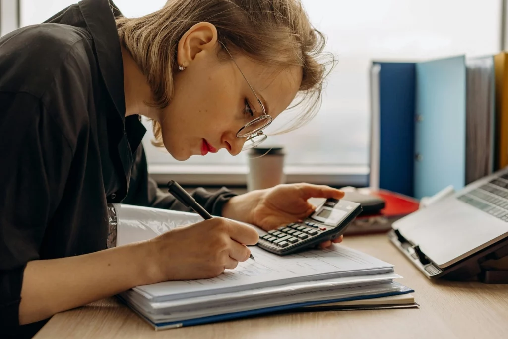 Mulher em uma mesa com documentos e uma calculadora, enquanto olha minuciosamente o documento.